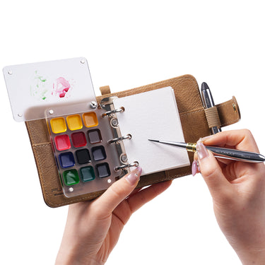 Silver Fuumuui Round travel watercolor brush painting on cold press 100% cotton paper in a mini sketchbook. Features a 12-well travel palette and detail brush in a leather binder for professional artists.