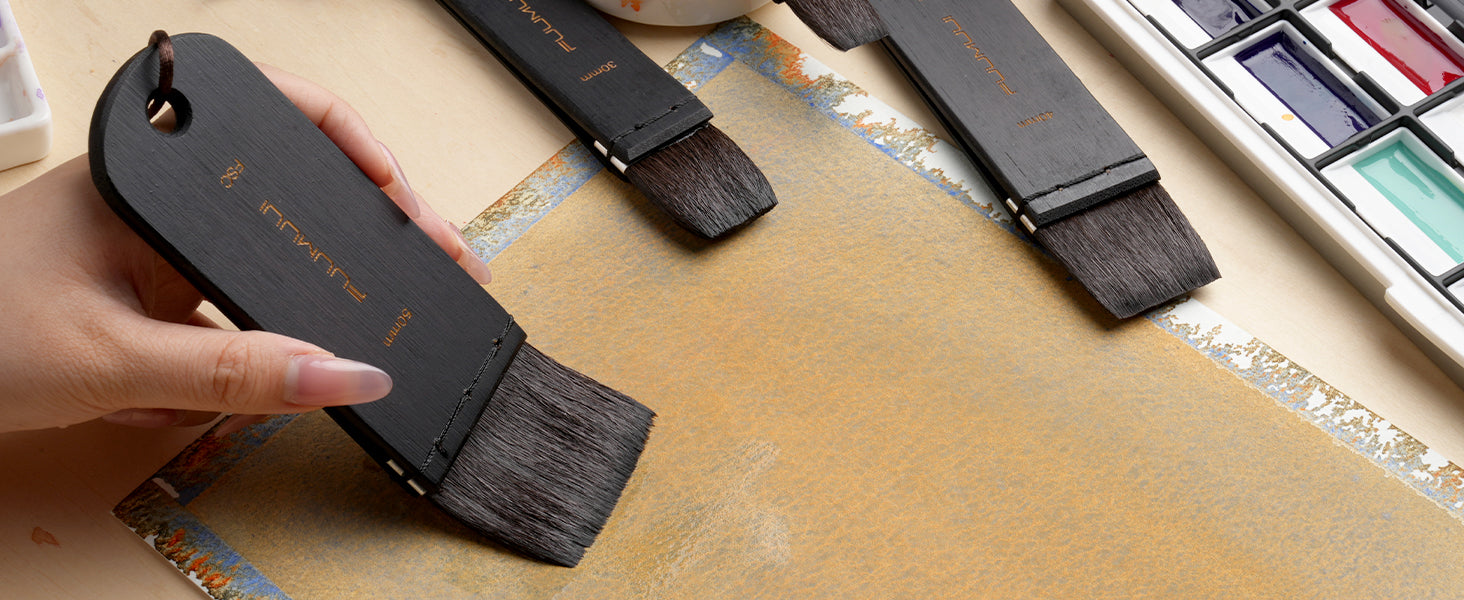 Fuumuui 50mm Squirrel Hair Wash Brush being used on watercolor paper by a professional artist. 30mm and 40mm flat brushes and a watercolor paint set are nearby, ideal for wide washes and smooth backgrounds.