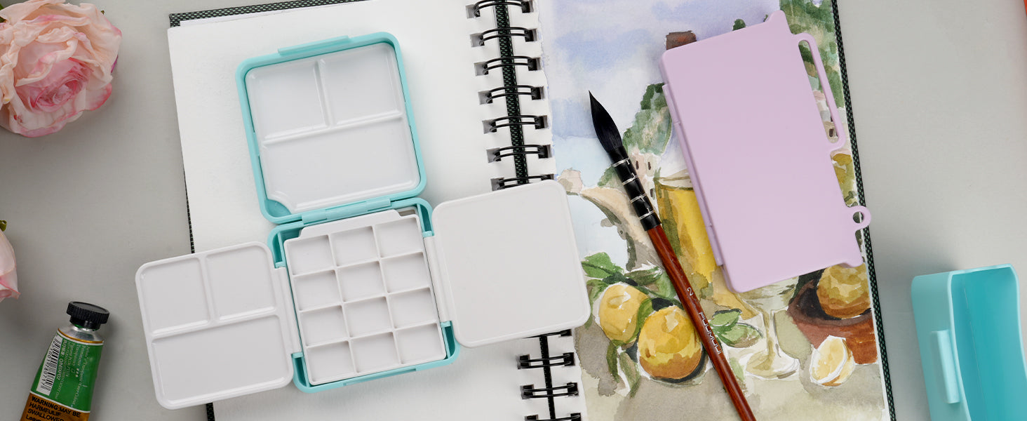 Fuumuui size 2 squirrel hair watercolor brush resting on a cold press sketchbook next to an open mint green plastic travel palette and watercolor paint tube; professional artist and beginner art supplies.