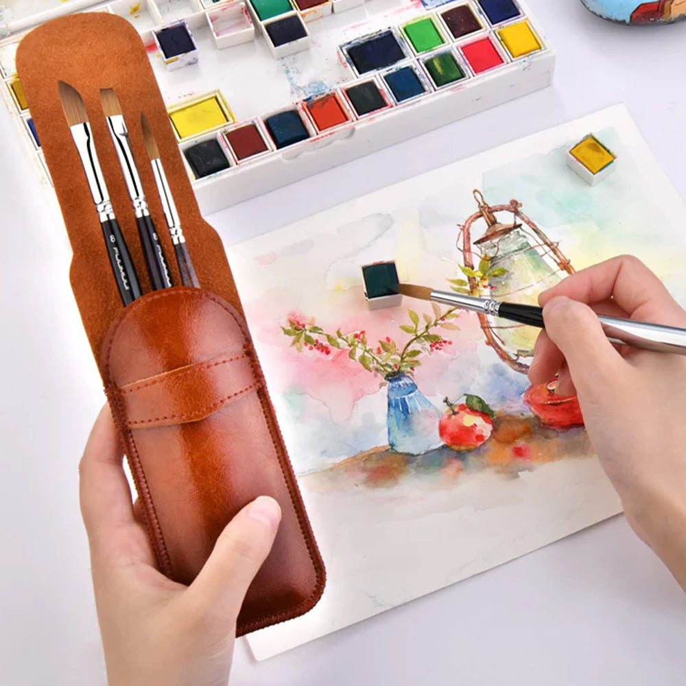 A hand using a Fuumuui sable travel watercolor brush to paint a landscape, with the leather case and a watercolor palette visible.