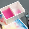Fuumuui Ceramic Paint Brush Wash Bowl with two compartments, one filled with pink water from rinsing and the other with clean water, positioned next to a watercolor painting and an artist's brush.