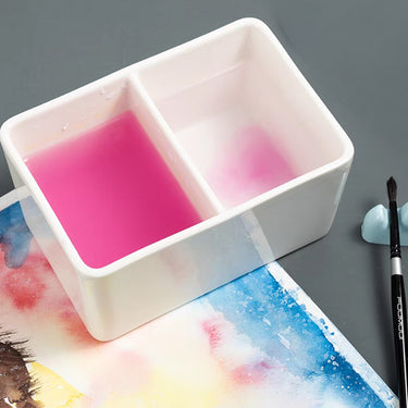 Fuumuui Ceramic Paint Brush Wash Bowl with two compartments, one filled with pink water from rinsing and the other with clean water, positioned next to a watercolor painting and an artist's brush.