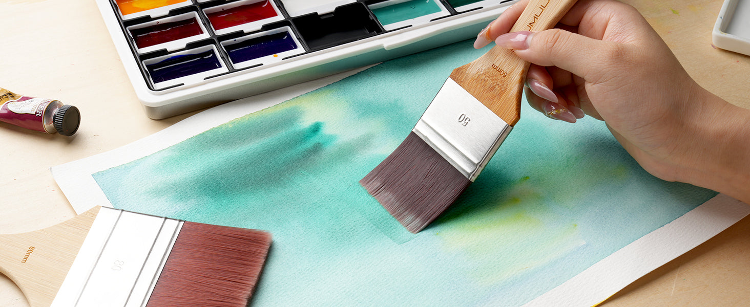 Professional artist using a 50mm Synthetic Nylon flat wash watercolor brush on textured cold press paper. Includes an 80mm flat brush and a watercolor paint set palette for creating smooth pigment washes and backgrounds.