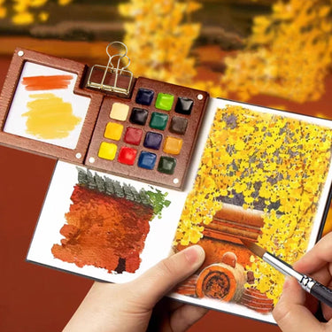 Mini-boîte de peinture aquarelle en noyer Fuumuui ouverte avec des godets d'aquarelle vibrants, une main peignant un paysage d'automne et des échantillons de couleurs sur la palette de mélange.