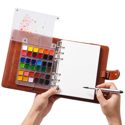 Portable watercolor paint set with a translucent travel palette and silver detail brush. Hands holding a brown leather sketchbook with 100% cotton cold press paper, ideal for professional artists and beginners.