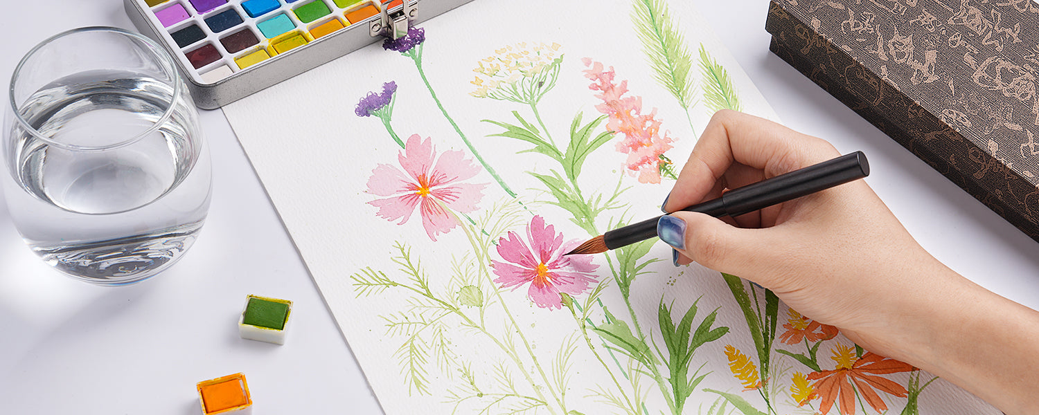 Professional artist using a Kolinsky Sable Travel Watercolor Brush to paint pink flowers on 100% cotton Cold Press paper. A metal tin Watercolor Paint Set and water jar are nearby on the table.