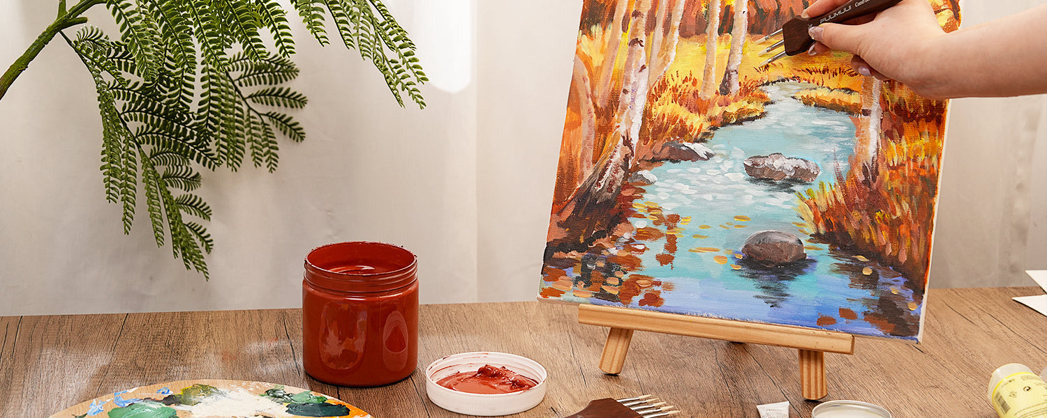 Artist using a Fuumuui Synthetic Nylon Comber brush to paint an autumn landscape with Acrylic Paint. Professional art supplies including a plastic palette and red paint jar on a wooden table.