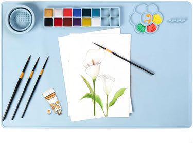 Tapis de peinture en silicone FUUMUUI avec gobelet de lavage de pinceaux et palettes d'art intégrés, contenant des pinceaux et une œuvre d'art florale à l'aquarelle sur une surface bleu clair.