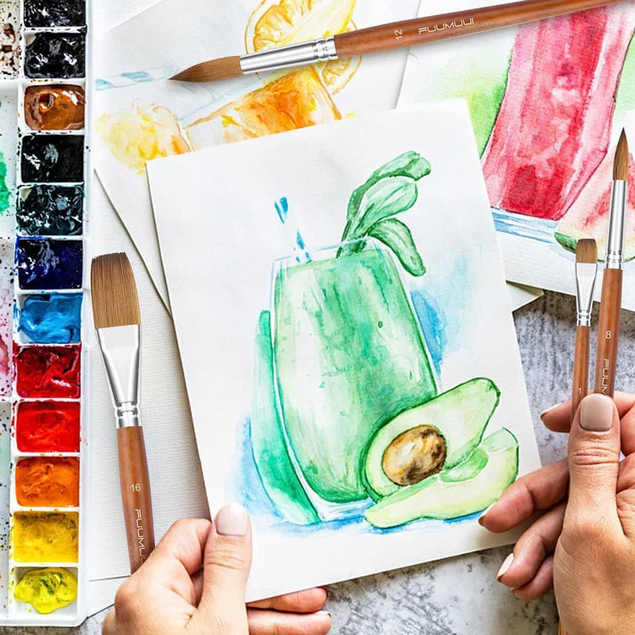 An artist holding a Fuumuui watercolor brush while painting an avocado, surrounded by a palette and art supplies for creative projects.