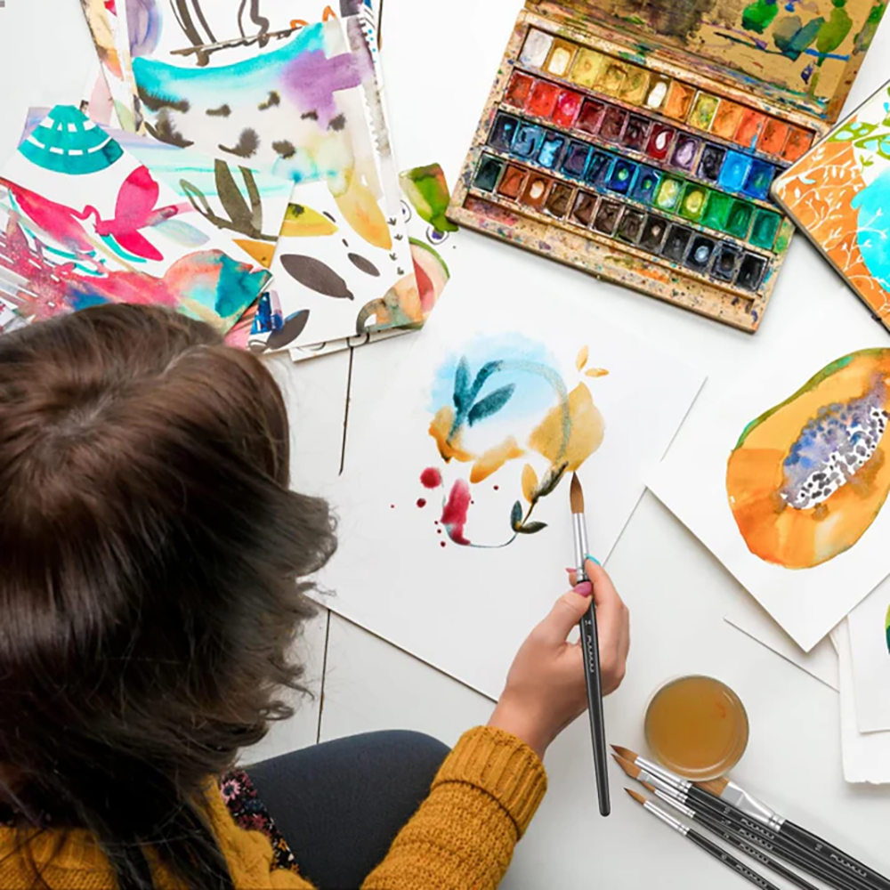 An artist uses a Fuumuui professional sable watercolor brush for painting, surrounded by art supplies and sketches.
