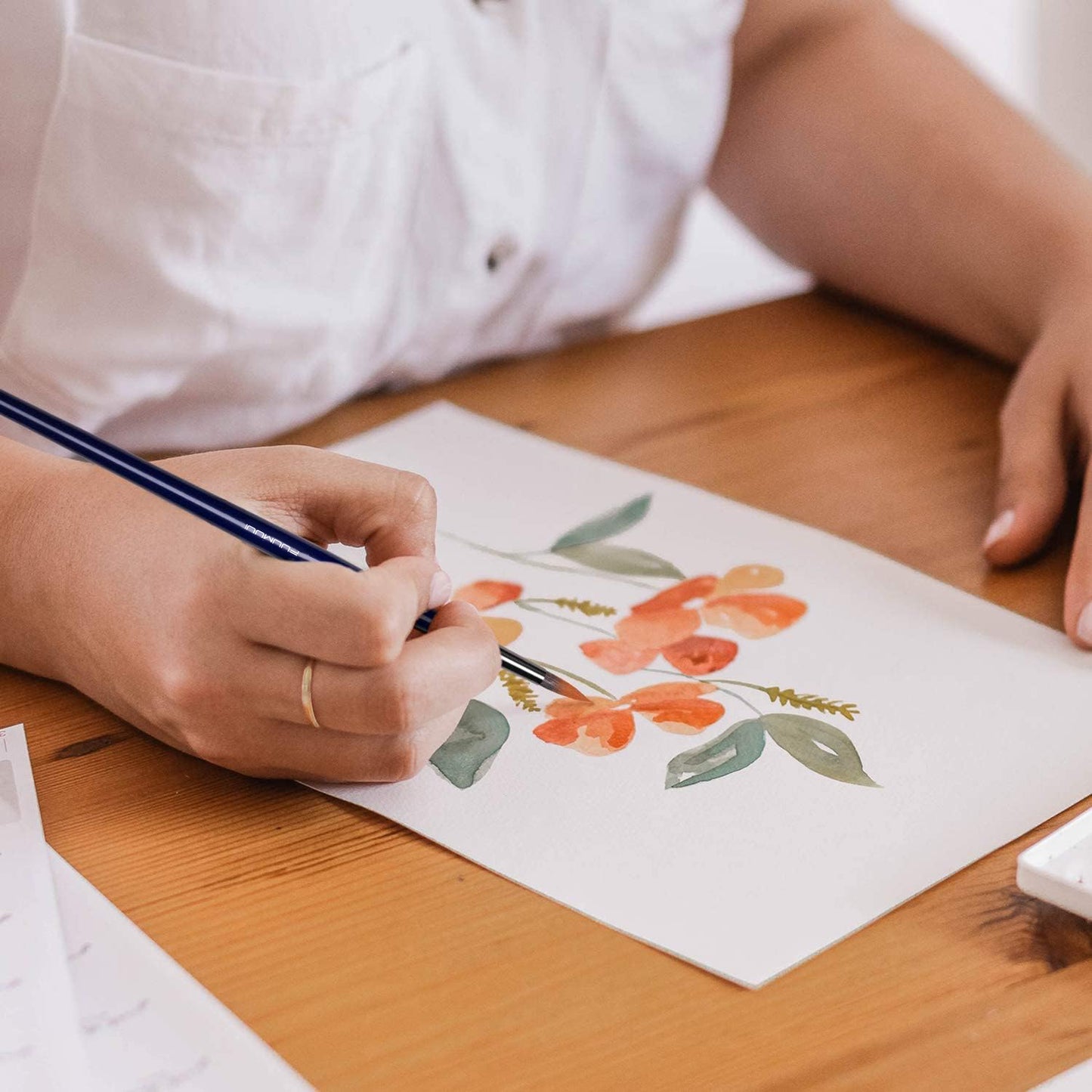 A person's hands using a Fuumuui professional sable watercolor brush to paint a floral pattern on paper.