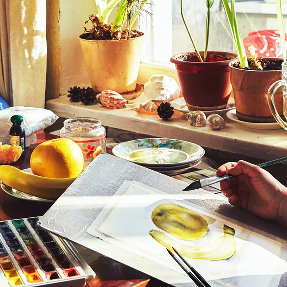 An artist's hand using a watercolor brush to paint a banana, with art supplies, fruit, and plants visible in the background.