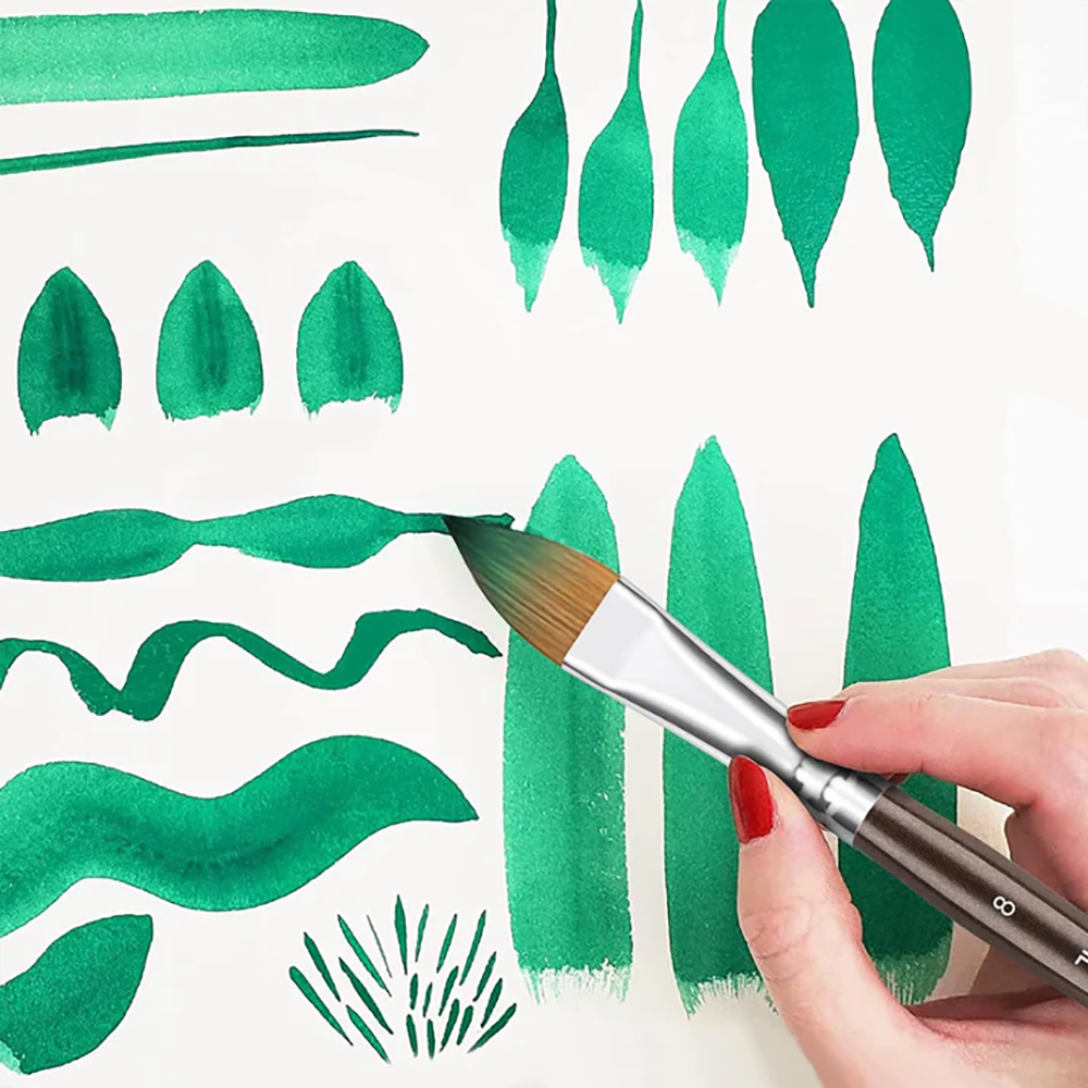 A hand holding a watercolor brush creating different green strokes and leaf shapes on white paper.