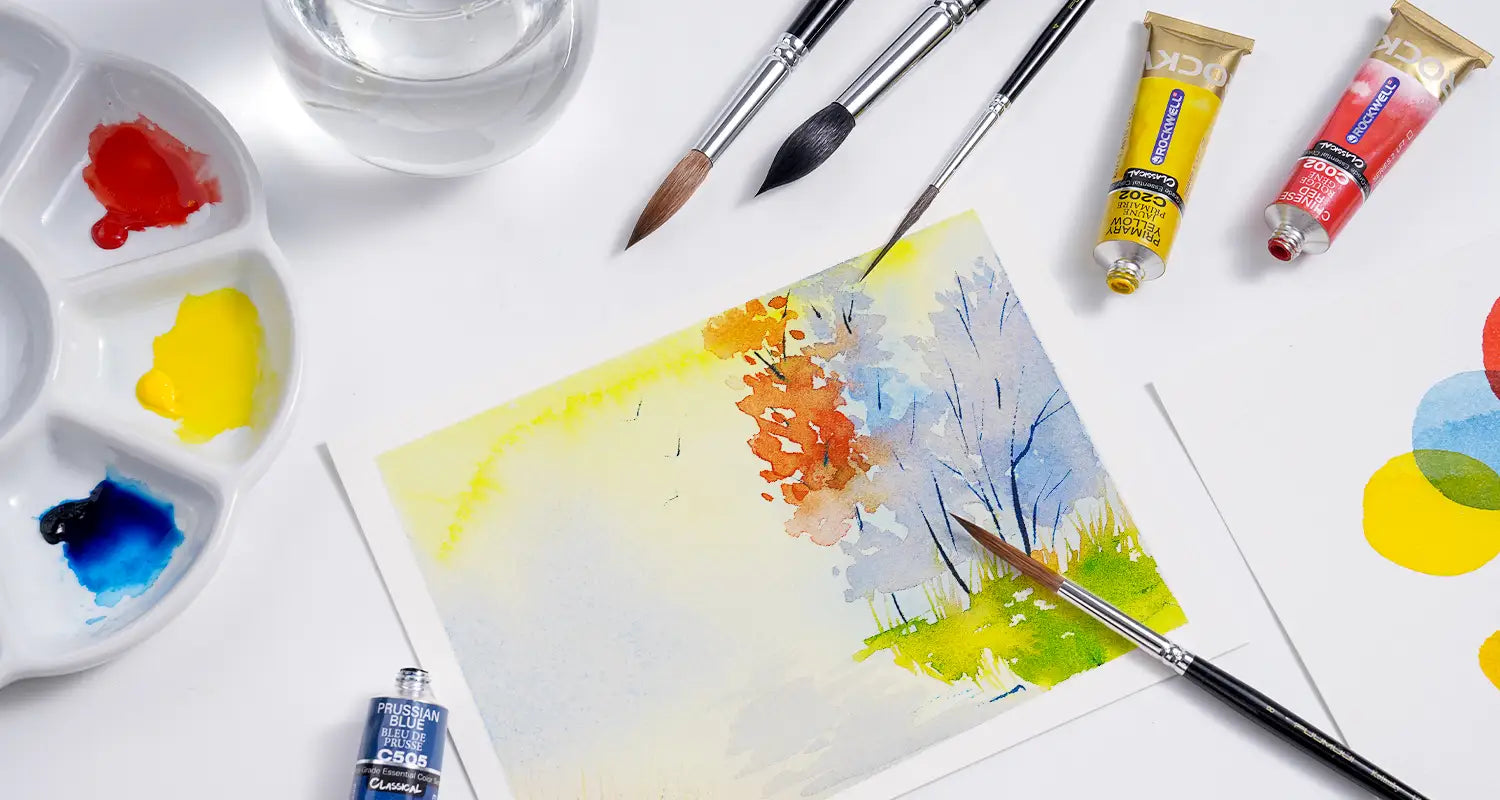 Watercolor artwork in progress surrounded by FUUMUUI brushes, ROCKWELL paint tubes, a water jar, and a palette.