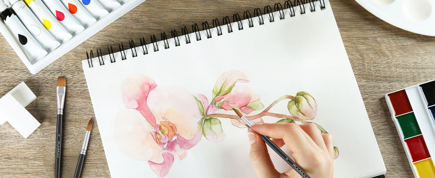 Professional artist uses a Fuumuui size 4 Kolinsky Sable watercolor brush to paint in a cold press sketchbook. A size 8 flat brush and watercolor paint set with tubes are organized on a wooden desk.