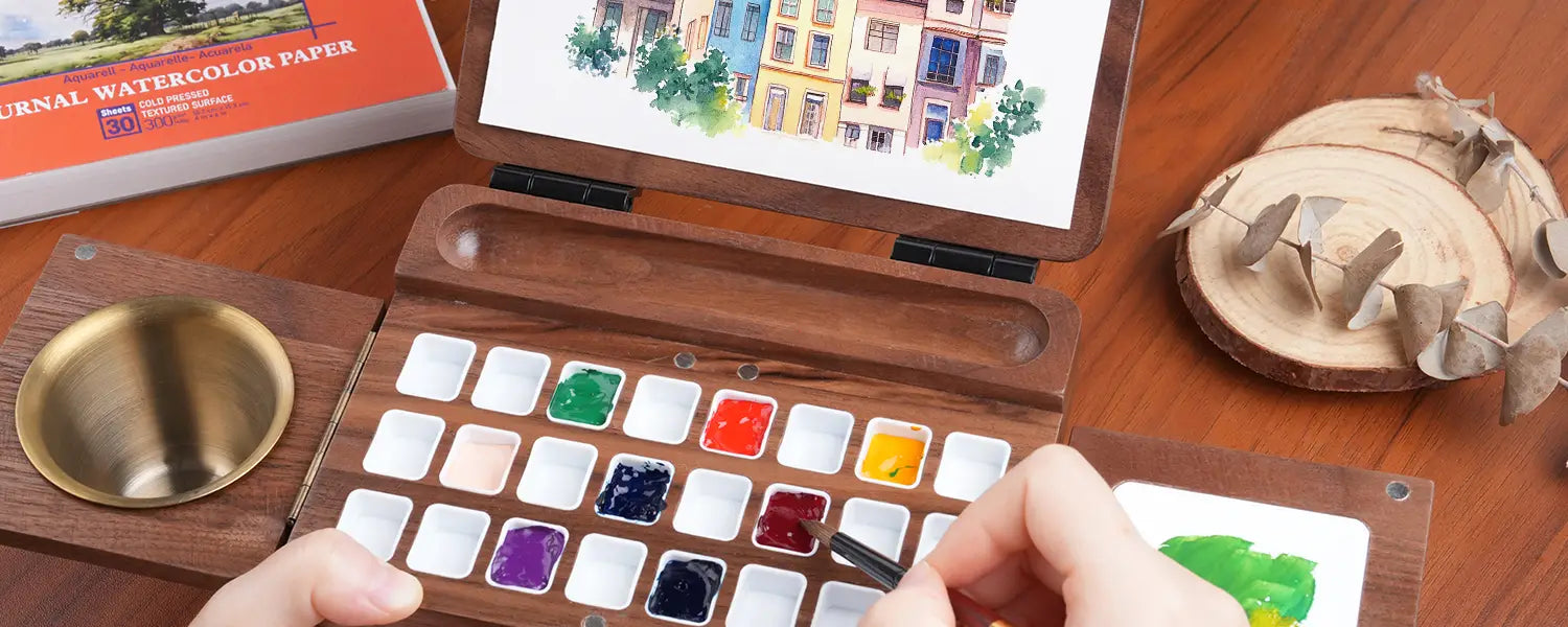 Hand using a sable travel watercolor brush with a 24-grid foldable wooden travel palette, professional cold press 300gsm watercolor paper journal, and brass brush cleaner cup on a wooden workspace.