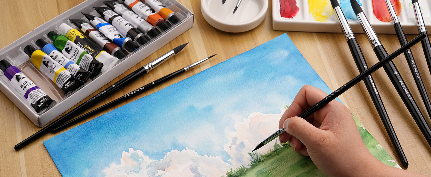 Professional artist using a Fuumuui synthetic squirrel hair round watercolor brush from a 9-piece set on cold press paper. Includes watercolor paint tubes, a ceramic palette, and assorted round brushes.