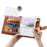 Artist painting a winter landscape in a large watercolor travel journal set with 36 semi-solid paints.