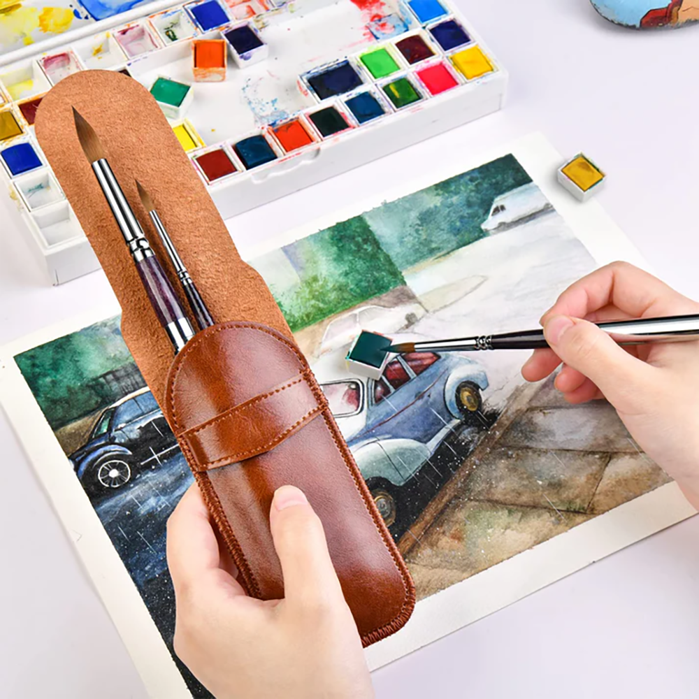 A hand using a travel watercolor brush to paint a car on paper, holding the kit's leather case.