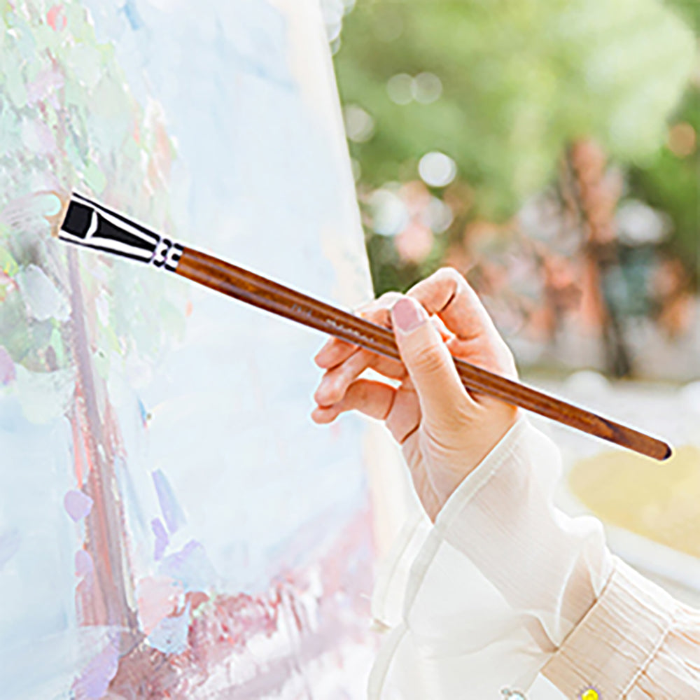 An artist holds a large flat hog bristle brush, painting outdoors with it on an easel.