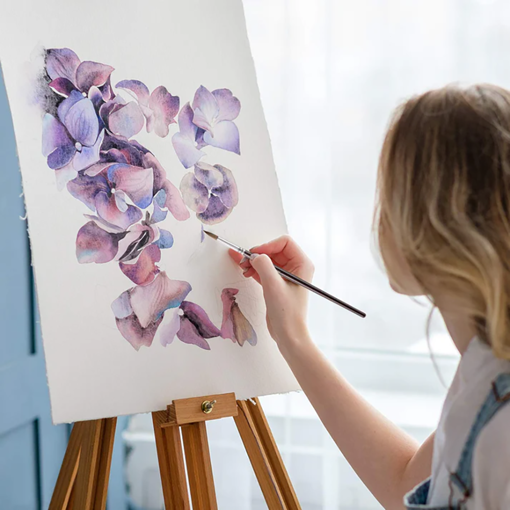 A painter uses a Fuumuui sable watercolor brush to create a beautiful floral painting on a canvas on an easel.