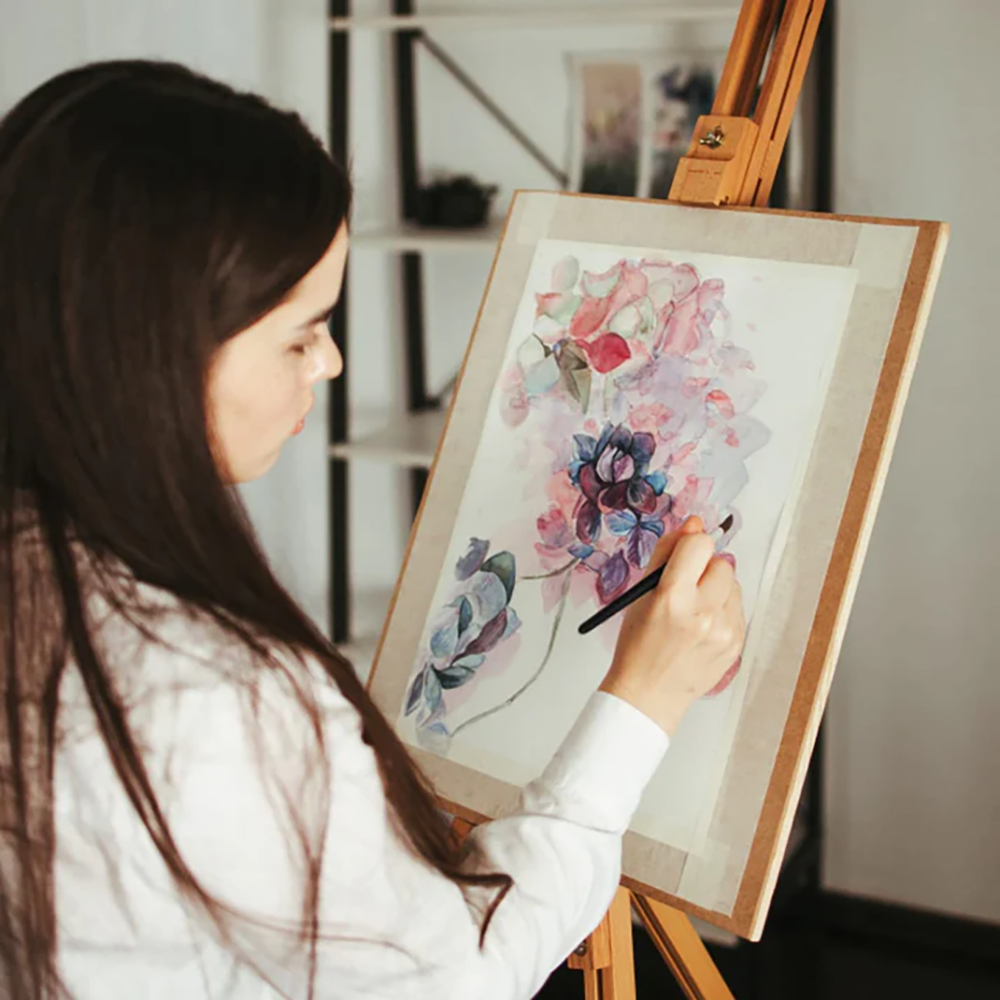 An artist uses a Fuumuui watercolor brush to paint a beautiful floral design on a canvas mounted on an easel.