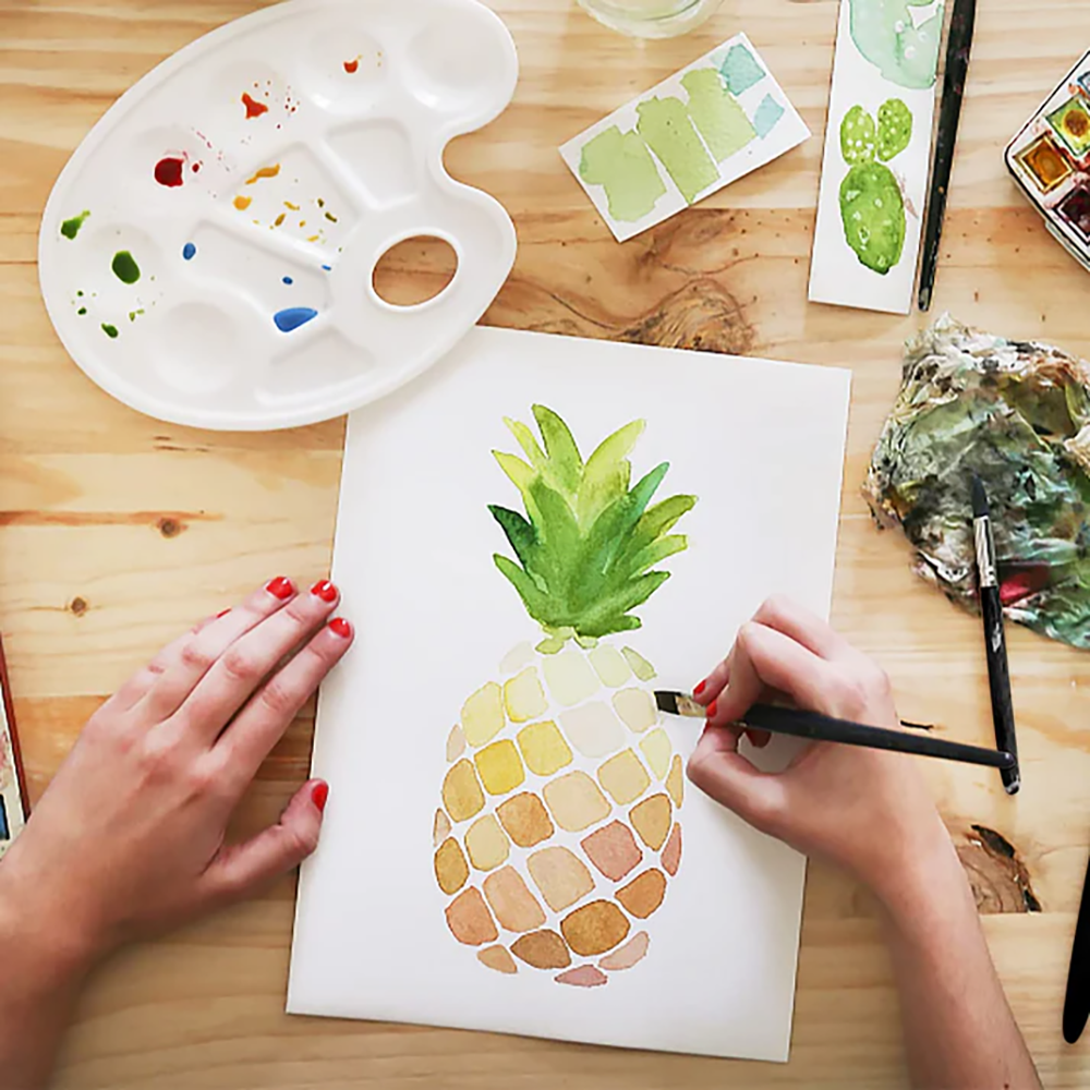 Fuumuui watercolor brushes in use by an artist painting a pineapple, surrounded by a palette and other art supplies.