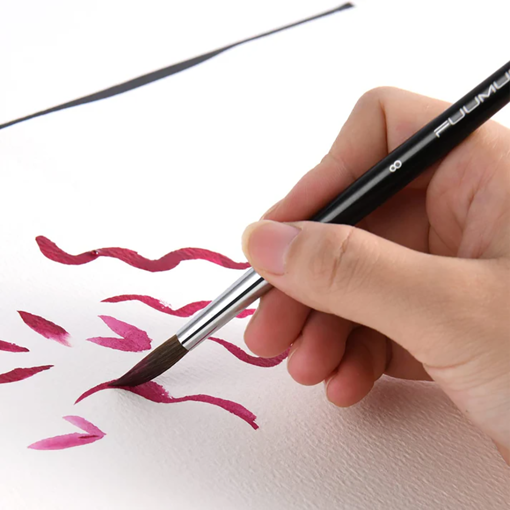 A hand holds a Fuumuui round watercolor brush, creating wavy red and pink strokes on a white paper surface.