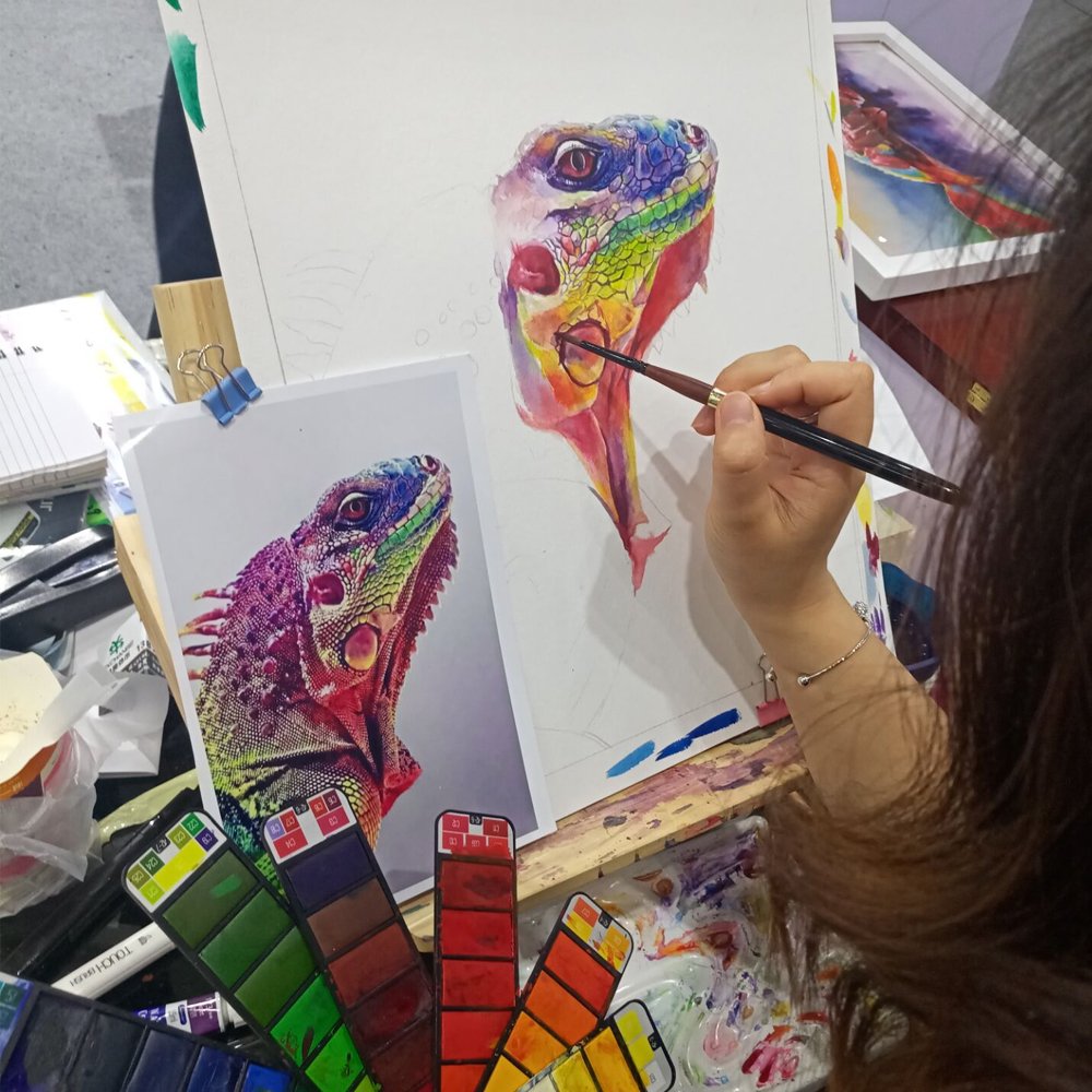 A person uses a professional watercolor brush set to paint a colorful iguana, with the reference photo and a color swatch card on the easel.