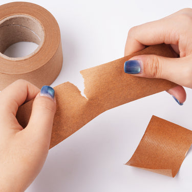 Hands tearing Fuumuui Watercolor Stretching Kraft Water-Activated Paper Tape, revealing its textured adhesive surface, with a roll of Fuumuui 45mm art supplies tape in the background.