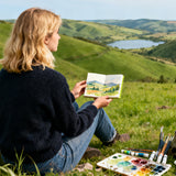 Una donna usa il suo blocco per acquerello Fuumuui, un album da schizzi 5.5x5.5" Verde Erba, per dipingere un bellissimo paesaggio all'aperto. ¨¨ seduta su una collina erbosa con la sua carta 100% cotone, pressata a freddo, aperta, dipingendo le dolci colline verdi e il lago distante. Vari materiali artistici Fuumuui, tra cui vernici e pennelli, sono disposti accanto a lei.