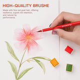 Una mano tiene un pennello per acquerello in pelo di capra rosso FUUMUUI, realizzato a mano, con manico rosso, dipingendo un delicato fiore rosa su carta, mostrando il suo eccellente assorbimento e flusso. Tre godet di acquerello di materiali artistici FUUMUUI (rosso, arancione, verde) sono visibili nelle vicinanze.