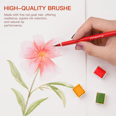 Una mano tiene un pennello per acquerello in pelo di capra rosso FUUMUUI, realizzato a mano, con manico rosso, dipingendo un delicato fiore rosa su carta, mostrando il suo eccellente assorbimento e flusso. Tre godet di acquerello di materiali artistici FUUMUUI (rosso, arancione, verde) sono visibili nelle vicinanze.
