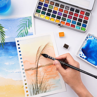 Une main utilise un pinceau aquarelle professionnel Fuumuui avec corps imprimés pour peindre un paysage de coucher de soleil, avec un ensemble de peinture aquarelle Fuumuui visible en arrière-plan.