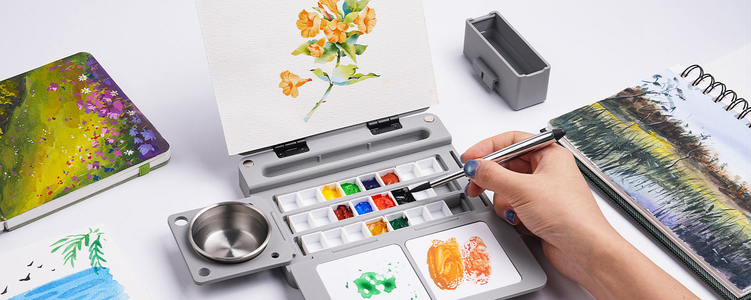Professional artist using a silver Travel Watercolor Brush and portable Travel Palette watercolor paint set to paint flowers on a Cold Press paper sketchbook with an attached metal brush cleaner cup.