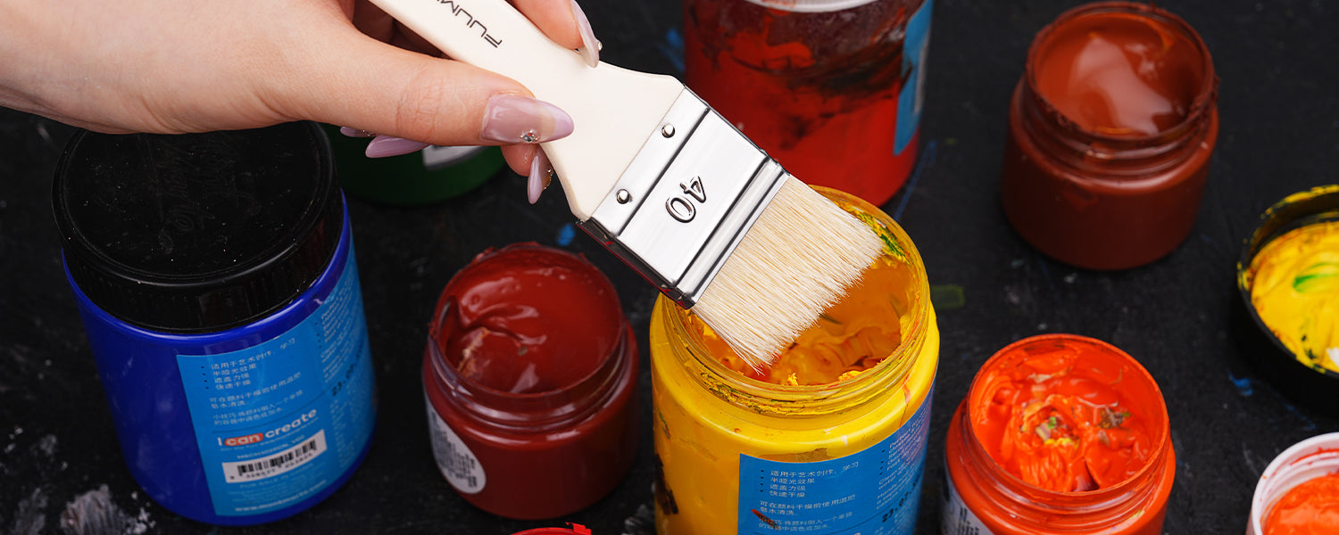 Fuumuui size 40 flat acrylic paint brush with white synthetic nylon bristles dipping into a jar of yellow acrylic paint pigment. Professional artist tool for wide coverage and large scale painting.