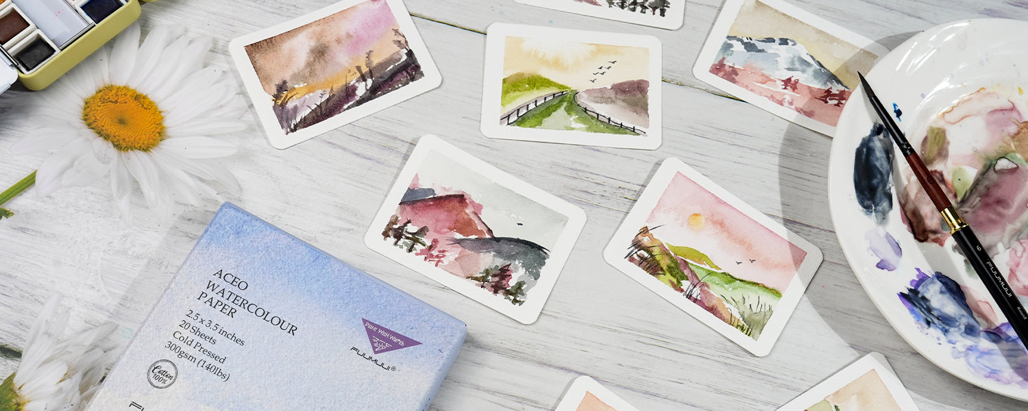 ACEO watercolor paper set with painted cards, brush, palette, and box arranged on a white floral surface.