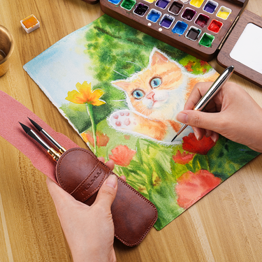 Fuumuui Art Supplies: Artist painting a watercolor cat illustration using a Fuumuui 3pcs Small Dual-Tip Silver Travel Watercolor Brush with Premium Synthetic Squirrel Hair, while holding the brown Fuumuui brush case for 6 tip styles, next to a wooden watercolor palette.