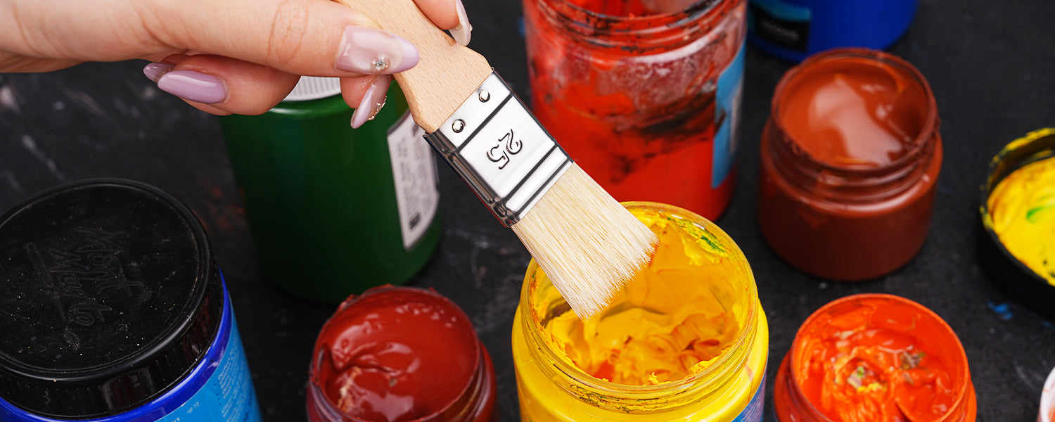 Artist dipping a Size 25 natural bristle flat brush into a jar of vibrant yellow acrylic paint. Multiple open jars of non-toxic pigments for professional artists and beginners to use for acrylic or oil paint.