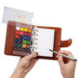 Fuumuui Watercolor Travel Set open, showcasing its brown PU leather cover, refillable ring-bound 300gsm cotton paper, vibrant premium watercolor paints, and clear mixing palette with swatches. A hand holds the double-ended travel brush with carbon lead, highlighting this portable Fuumuui art supplies kit.