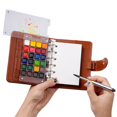 Fuumuui Watercolor Travel Set open, showcasing its brown PU leather cover, refillable ring-bound 300gsm cotton paper, vibrant premium watercolor paints, and clear mixing palette with swatches. A hand holds the double-ended travel brush with carbon lead, highlighting this portable Fuumuui art supplies kit.