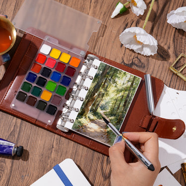 Fuumuui Watercolor Travel Set featuring a PU leather cover, open to reveal premium watercolor paints and refillable ring-bound 300gsm cotton paper, with an artist using the Fuumuui double-ended travel brush to paint a forest scene.