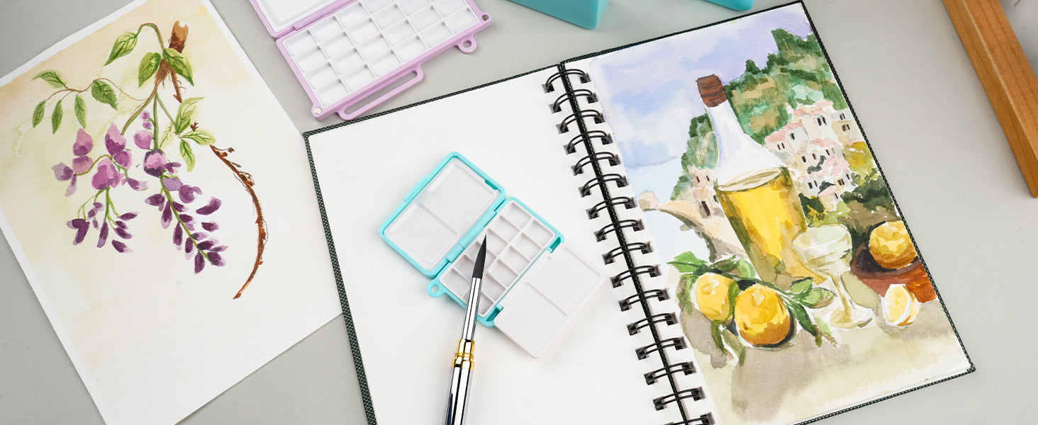 Synthetic nylon travel pocket brush resting on a plastic travel palette inside a spiral sketchbook with cold press paper, featuring professional artist floral and landscape watercolor paintings.