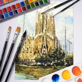 Ensemble de pinceaux de mélange aquarelle professionnels Fuumuui & Allan Jay Quesada avec des pinceaux plats, ronds et en forme de dague affichés autour d'une peinture aquarelle vibrante d'une cathédrale, à côté d'une palette de peintures colorées.