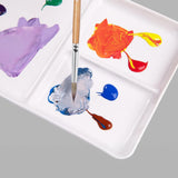 Palette de peinture d'artiste en céramique Fuumuui en cours d'utilisation, présentant des peintures violettes, orange, bleues et brunes vibrantes mélangées dans ses multiples godets en céramique blanche par un pinceau.