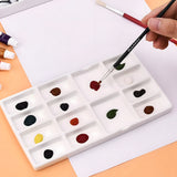 Palette de peinture en céramique pour artiste Fuumuui avec plusieurs godets remplis de diverses couleurs de peinture, une main tenant un pinceau Fuumuui trempant dans de la peinture rouge.