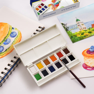 Fuumuui Tragbares 12-Farben Festes Aquarellfarben Set mit integrierter Palette und Pinsel, offen auf einer weißen Oberfläche neben leuchtenden Fuumuui Art Supplies Aquarellmalereien und einem Skizzenbuch.