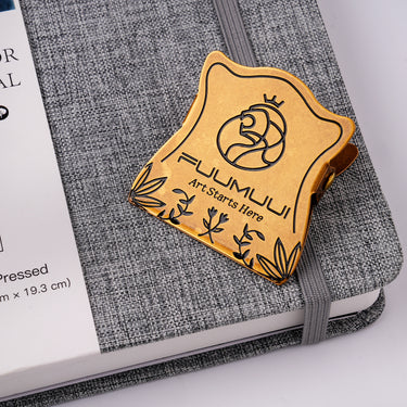 Fuumuui vintage brass clip with intricate lion logo and leaf engravings, displayed on a grey textured sketchbook, ideal for artists using watercolor paper.