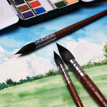 Set di pennelli a punta tonda in pelo di scoiattolo naturale FUUMUUI da 6 pezzi per arte ad acquerello, con tre pennelli con manico scuro su un dipinto paesaggistico scenico, con una tavolozza di acquerelli di forniture artistiche FUUMUUI sullo sfondo.