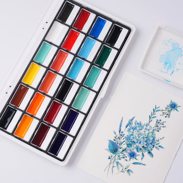 Set di colori ad acquerello opachi semisolidi a 24 colori Fuumuui - Colore terra, mostrato con un vivace dipinto ad acquerello floreale blu e una tavolozza di miscelazione.